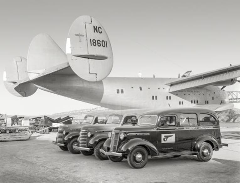 Black and white 1939 photo of the Honolulu Clipper airplane behind three black automobiles