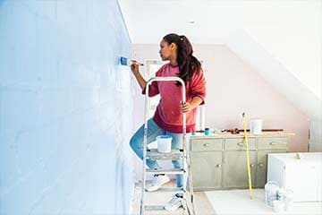 Woman paints a wall in her home while standing on a small ladder.