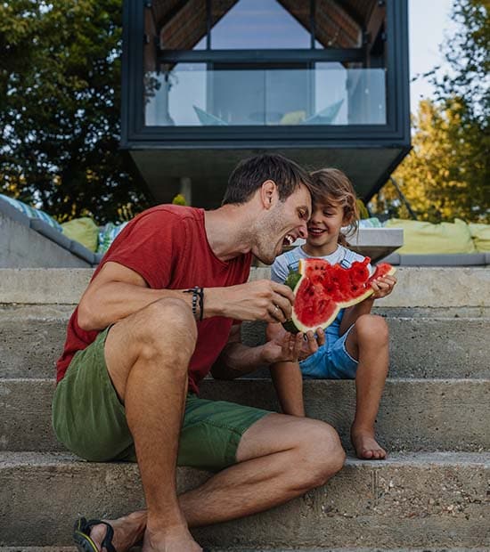 A father sits with his child on steps behind their home. They eat watermelon together.