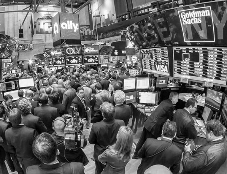 Black and white 2014 photo of a crowded stock exchange floor, with Ally logos appearing on overhead monitors