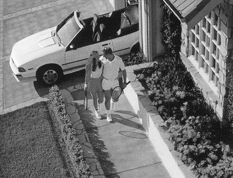 Black and white 1989 print ad photo of a white male and female wearing tennis gear and holding tennis rackets as they walk from their white convertible car to their house