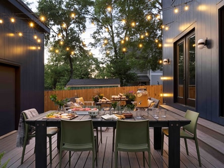 A backyard patio is decorated with string lights above a dining table. The table is set for an evening of entertaining.
