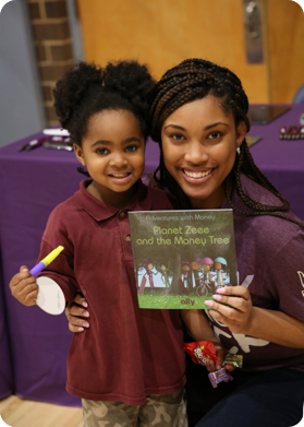 One of Ally’s Black teammates posing next to a young Black girl while holding up Planet Zeee and the Money Tree; a book that teaches financial literacy to children.