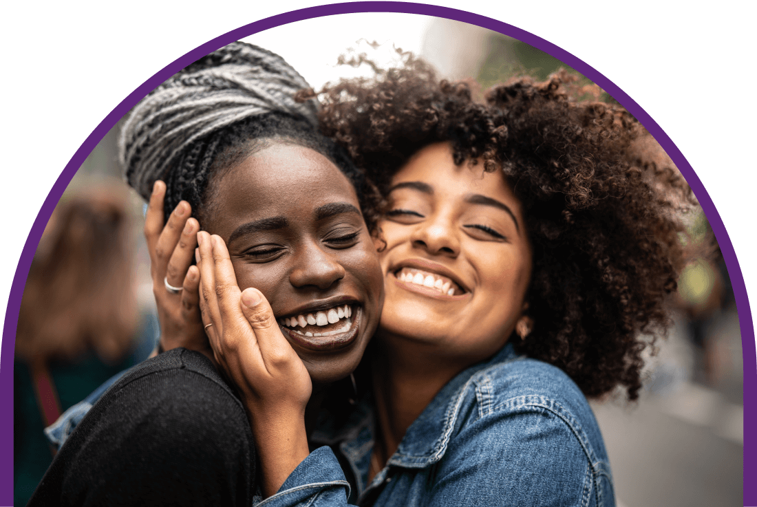 A caramel skinned woman smiles with her cheek pressed to the cheek of a smiling, ebony skinned woman.