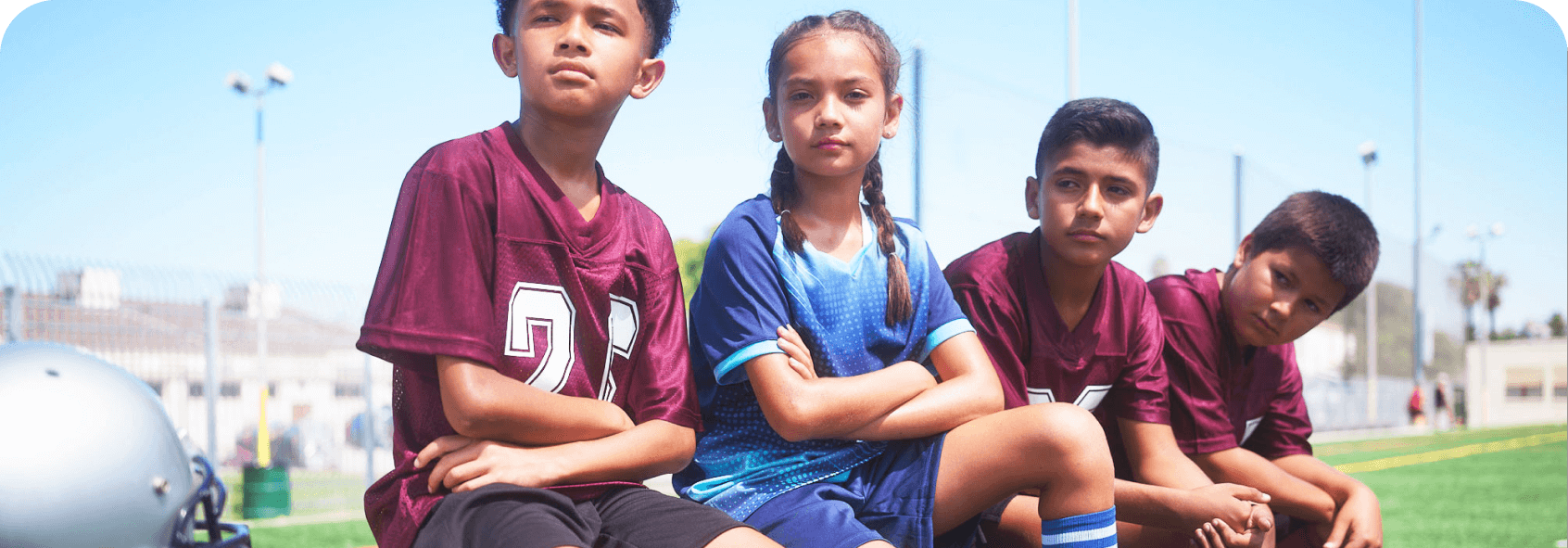 A group of Hispanic and Latino children wearing athletic gear and sitting on an athletic field.
