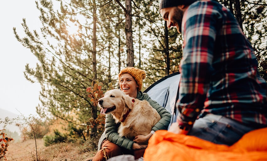 A smiling white woman wearing a beanie snuggles her golden retriever next to her car while her bearded white boyfriend sits nearby.