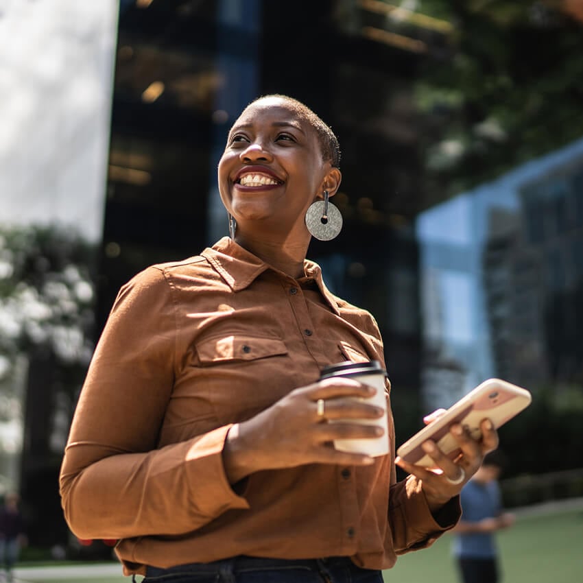 A smiling Black woman looks into the distance while holding a cup of coffee and her phone.