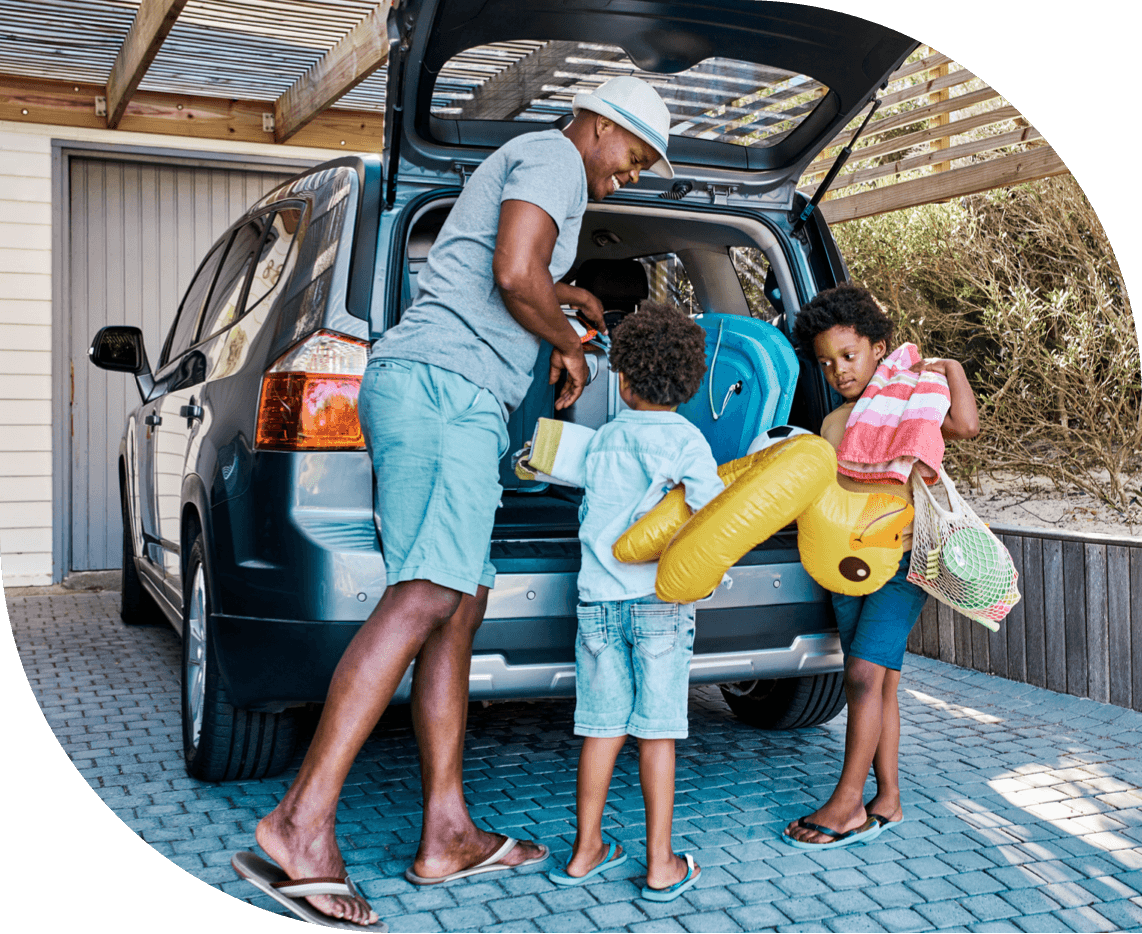 A smiling father and his two children dressed for the beach pack the children’s toys into the trunk of an SUV