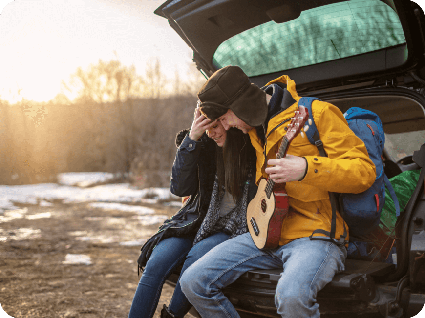 A smiling young couple sit together on their car’s tailgate dressed for a cold weather hike as one of them holds a ukelele.