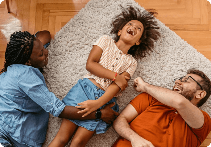 A young girl laughing while laying down on a rug with her parents.