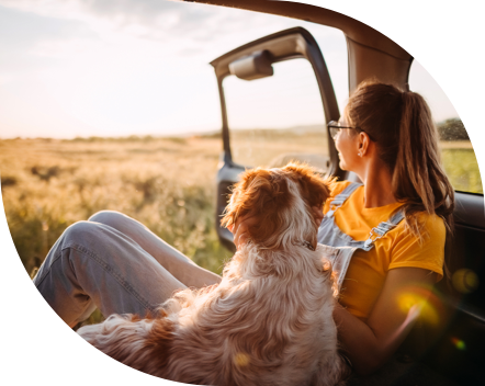 Image of a person with glasses and a ponytail wearing a yellow t-shirt and overalls sitting inside a vehicle next to a dog, both looking outside from the open door