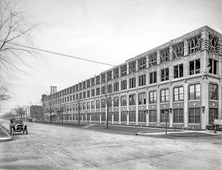 Black and white 1910 photo of the exterior of the Detroit Car Factory, with one black 1910 automobile in front
