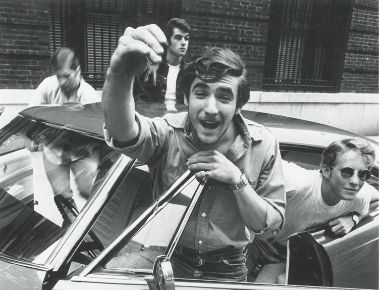 Black and white 1967 print ad photo of four white males wearing 60s fashion standing around an automobile, with the one by the driver’s side proudly holding up keys