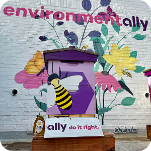 A mural of a bee hovering around a pot of flowers, with the words environmentally and ally do it right painted on a brick wall