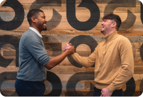 Two men shake hands in front of a wooden wall adorned with repeating lowercase A’s of the Ally logo.