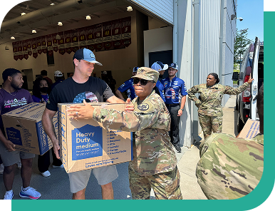 Outdoors at a donation event, an Ally volunteer passes a cardboard box full of donations to a person in a camouflage uniform.