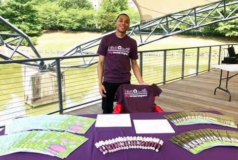 a young Ally employee at a community event wearing an Ally social impact T-shirt and standing behind a table with Ally merchandise on it.
