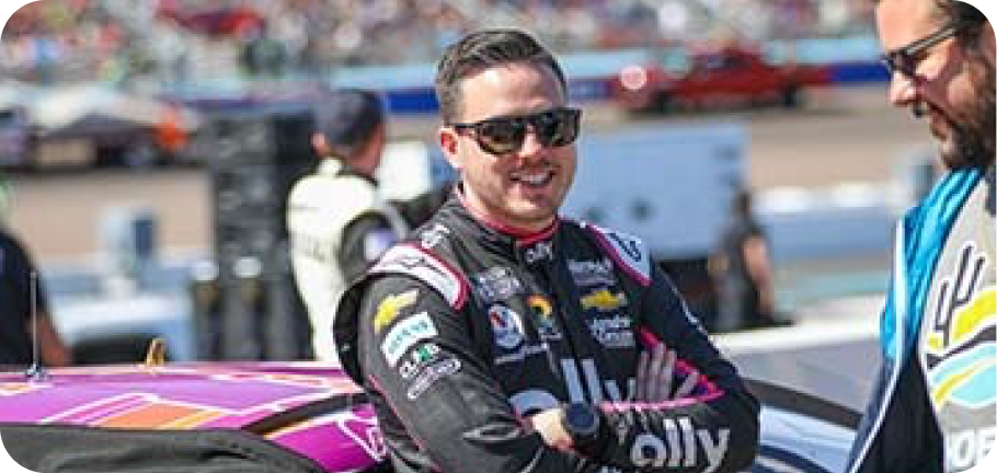 Alex Bowman leans against his NASCAR as smiles at a pit crew member.