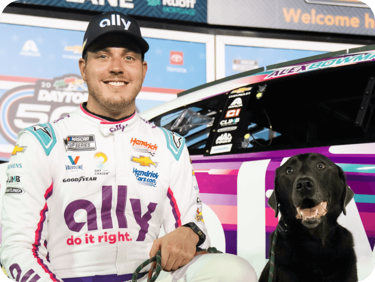 Alex Bowman poses with a black dog in front of the Ally racecar