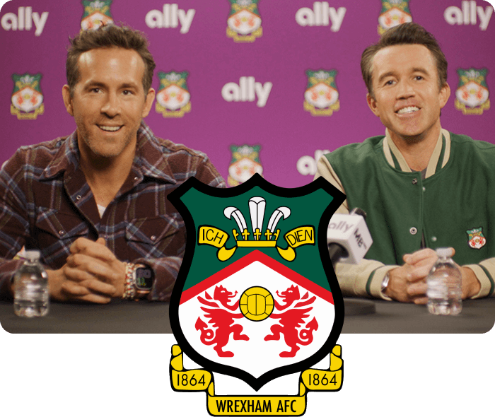 Rob McElhenney and Ryan Reynolds sit at a table behind a microphone in  front of a wall covered in Ally logos