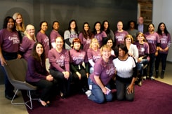 A large group of female Dress for Success participates posing in front of an Ally banner wearing matching purple Ally shirts