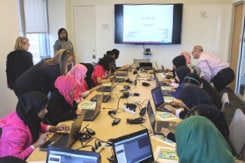 A group of young women sitting around a large table working on their laptops while Ally employees offer assistance