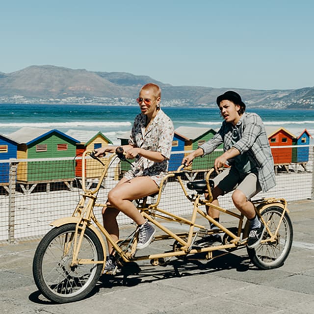 Two young people ride a tandem bike on a boardwalk by a shore with colorful houses and mountains in the background. 