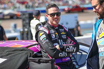 Alex Bowman leans against his NASCAR as smiles at a pit crew member.