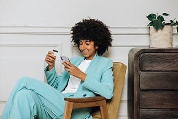 A woman is smiling while holding her phone and credit card in her hands to make an online purchase.