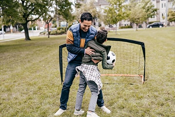 A happy father hugs his son while they play soccer in a park.
