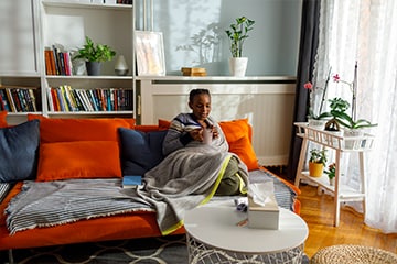 A woman with a cold sits on a couch with tissues and a hot drink