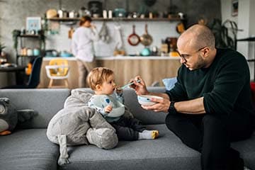 Father spoon feeding his infant son while his wife is in the background standing in the kitchen