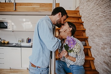 A man kisses a smiling woman on the nose while she sits on a staircase and holds flowers.
