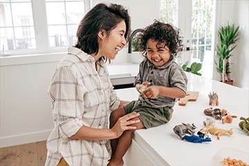  A mother and her toddler are smiling in a kitchen. The son's toys and some snacks are on the counter next to him.