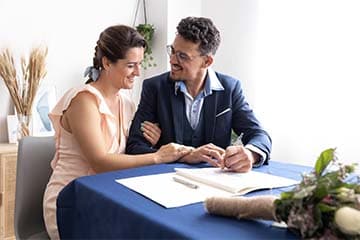 Couple getting married at home, sitting at a table signing the marriage certificate. 