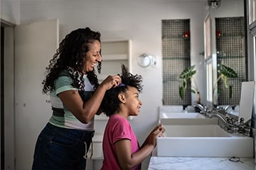 A mother smiles while combing her daughter's hair in a bathroom.