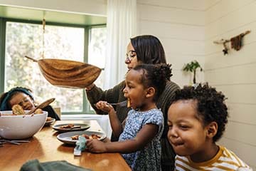 A mother is passing a salad bowl at a dining table while her three young children eat dinner. 