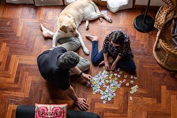 Two people sitting on the floor putting together a puzzle with their dog next to them watching