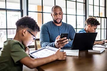 Father and two young sons sitting at a table with their tech. Son on the left has a tablet and son on the right has a laptop. The father is looking at his smartphone with his laptop open in front of him.