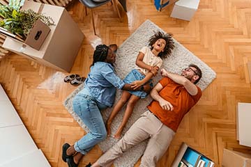 A mother, daughter and father are lying on the floor of their new dining room. They are surrounded by moving boxes.