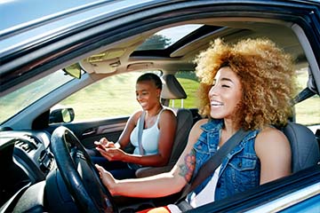Two women in a car laughing while driving with the windows down.