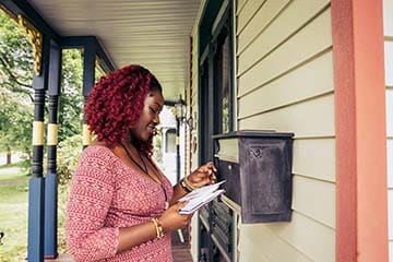 A woman is looking at her mail while standing on the porch of her home.
