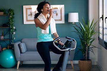 Young woman is drinking out of a water bottle while sitting on a stationary bike in her home.