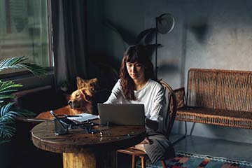 Young woman looking at her computer at her home. Her dog sits in a chair in the background.