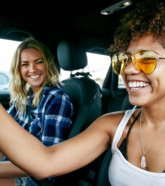 A curly-haired caramel-toned woman drives and laughs with her blonde-haired porcelain-toned friend.