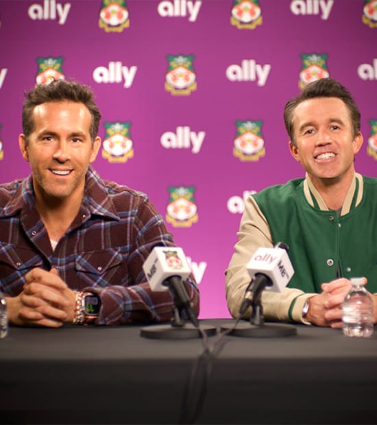 Ryan Reynolds and Rob McElhenney sit at a press conference table with Ally and Wrexham AFC logos on the wall behind them.