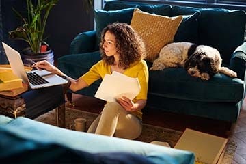 Woman on her laptop while holding some paperwork. Dog on the couch behind her.