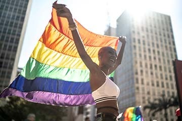A woman is smiling and holding a rainbow flag above her head while walking in a city.