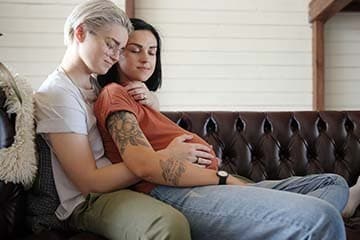 Young woman with fair hair hugs pregnant girlfriend with various tattoos sitting on brown leather sofa at home