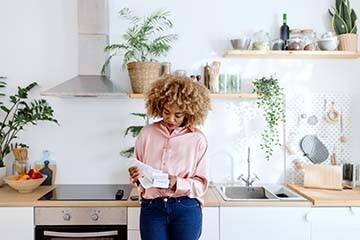A woman standing in her kitchen looking at receipts.